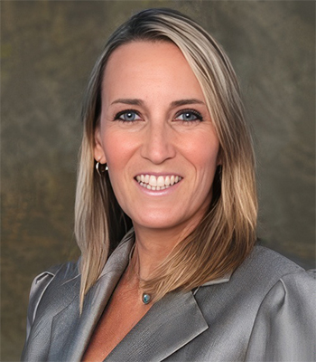 Smiling woman with straight blonde hair wearing a grey blazer, posing in front of a neutral brown background.