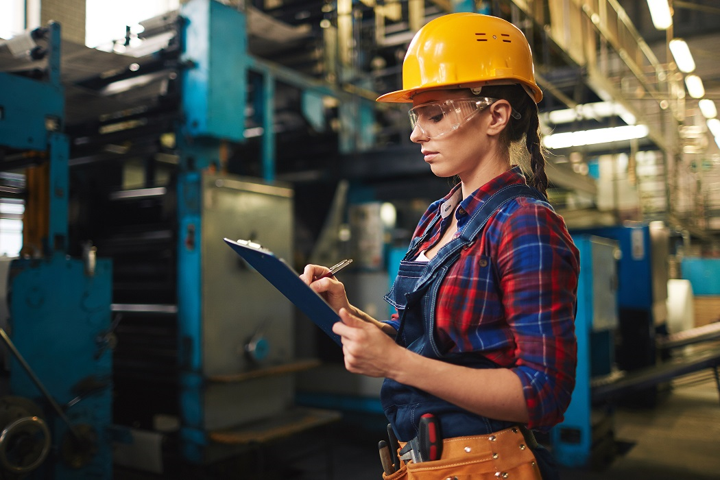 Woman in safety gear holding a clipboard and pen, inspecting equipment inside an industrial manufacturing facility.