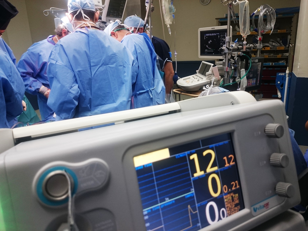 A group of surgeons in blue gowns operate in an operating room, with a heart rate monitor displaying vital signs in the foreground.