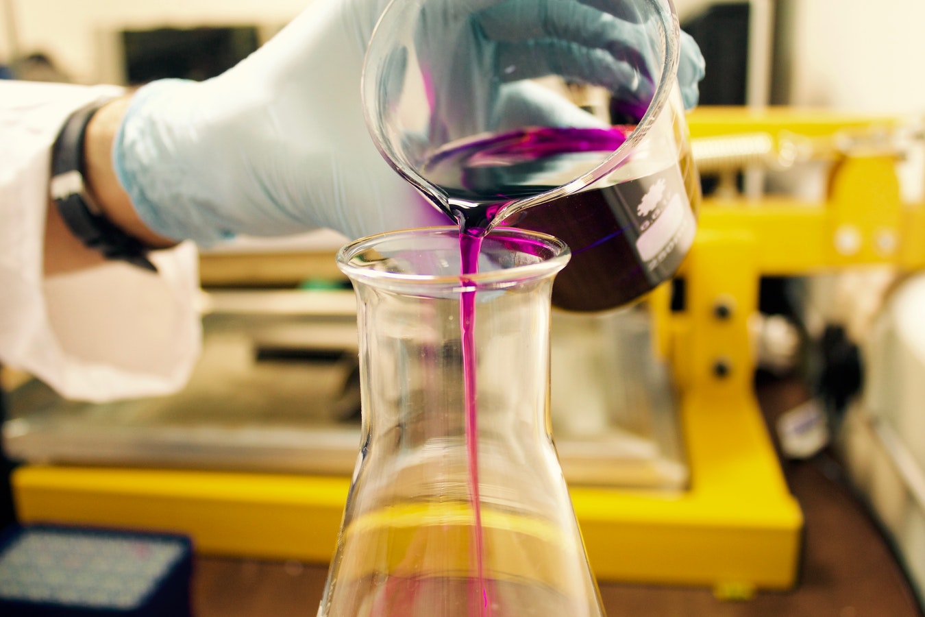 A gloved hand pours a purple liquid from a beaker into a clear Erlenmeyer flask in a laboratory setting.
