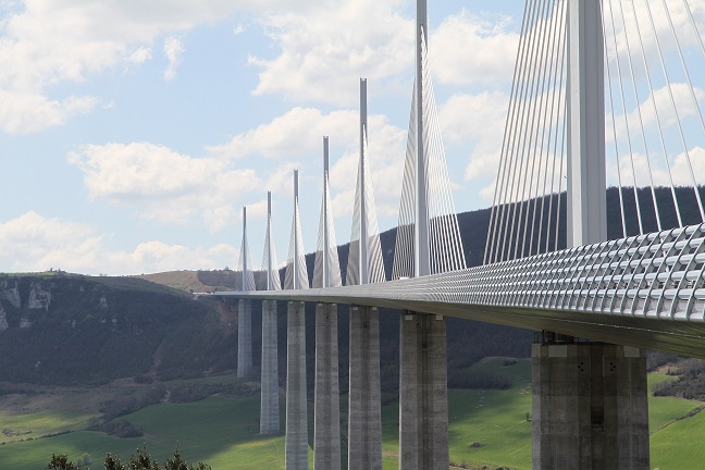 A large cable-stayed bridge with tall pylons spans a wide valley, with hills and grassy fields visible below.