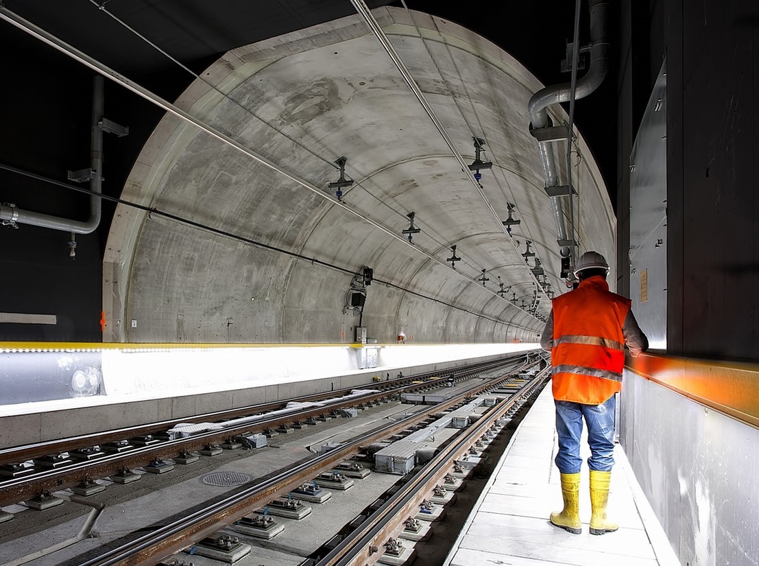 A worker in a reflective vest and yellow boots stands beside railway tracks inside a concrete tunnel, facing away from the camera.