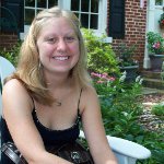A woman with blonde hair wearing a black top sits on a white chair outside, smiling, with greenery and a brick house in the background.