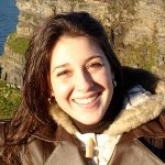 A woman with long brown hair wearing a brown coat with a fur-lined collar smiles outdoors with cliffs and water in the background.