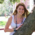 A woman with long brown hair, wearing a white sleeveless top and a necklace, stands outdoors beside a tree on a sunny day.