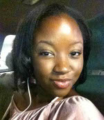 A woman with medium-length dark hair smiles slightly while sitting in a car, wearing a light pink top and white earphones.