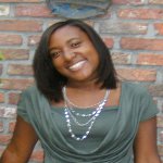 A woman with straight shoulder-length hair, wearing a green top and layered necklace, smiles while standing in front of a brick wall.