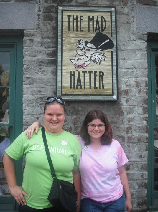 Two women stand smiling in front of a brick wall beneath a sign that reads "The Mad Hatter" with an illustrated hat-wearing animal.