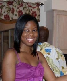A woman in a purple top smiles at the camera while seated indoors; a person in a floral shirt is visible in the background facing away.