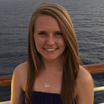 A woman with long straight hair smiles at the camera while standing on a boat deck, with the ocean in the background at sunset.