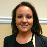 A woman with medium-length dark hair wearing a dark blazer and a black top stands indoors against a beige and white wall, smiling at the camera.
