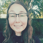 A person with long brown hair and glasses smiles outdoors, with trees and a wooden fence in the background.