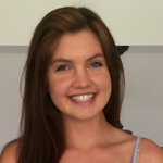 A young woman with long brown hair smiles at the camera. She is indoors and wearing a light-colored top.