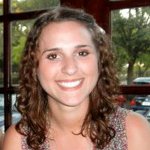 A young woman with curly brown hair smiles at the camera indoors, with large windows and trees visible in the background.