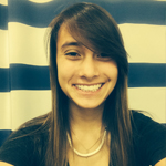 A young woman with long brown hair smiles at the camera, standing in front of a white and blue striped background.