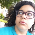 A person with curly hair and glasses looks off to the side outdoors, wearing a blue shirt, with trees and buildings in the background.