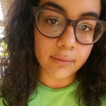 A person with long curly hair and glasses wearing a green shirt looks at the camera with a neutral expression.