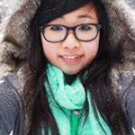 Young person with long dark hair, wearing glasses, a green scarf, and a hooded winter coat, standing outdoors in snowy surroundings, smiling at the camera.