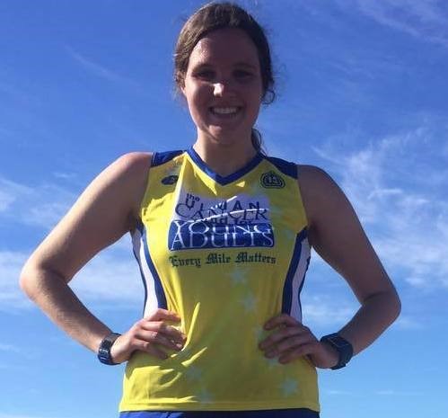 A woman stands outdoors under a blue sky, wearing a yellow athletic tank top with "Every Mile Matters" and "Iowa Abilities" written on it. She is smiling and has her hands on her hips.