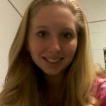 A young woman with long blonde hair is smiling at the camera indoors, with a plain wall and some shelves in the background.