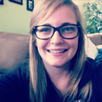 A young person with long blonde hair and black glasses smiles while sitting on a couch indoors.