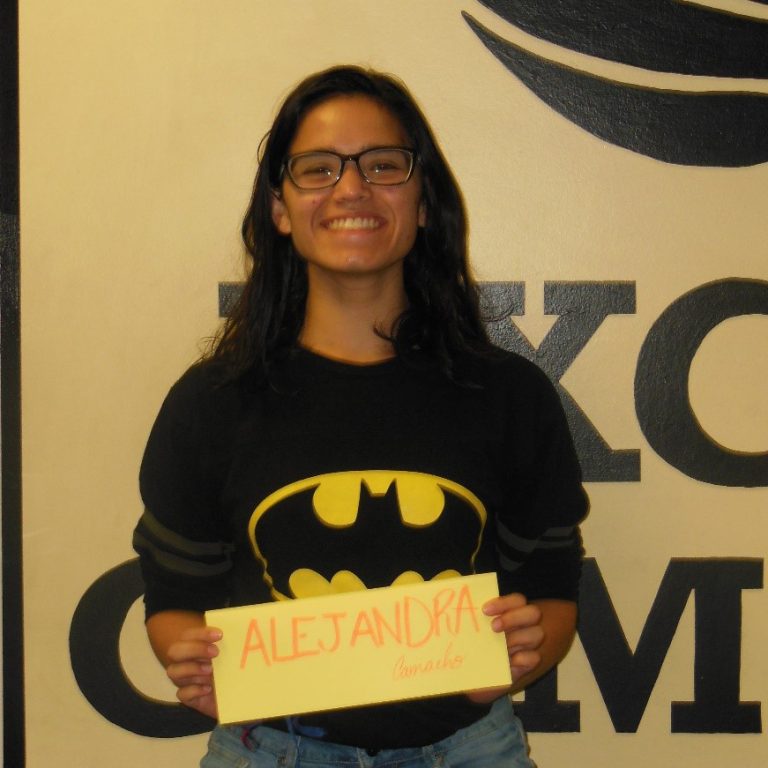 A person wearing glasses and a Batman logo shirt holds a yellow sign that says "Alejandra" in front of a wall with large black letters.