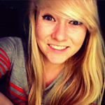 Young woman with long blonde hair and a gray shirt with red stripes smiles at the camera against a dark background.