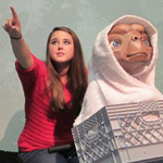 A young woman in a red shirt points to the left while sitting next to a figure of E.T. wrapped in a white blanket inside a crate.