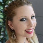 A young woman with long blonde hair and blue eyes smiles at the camera. She is wearing dangling earrings and bright red lipstick. Green plants are visible in the background.