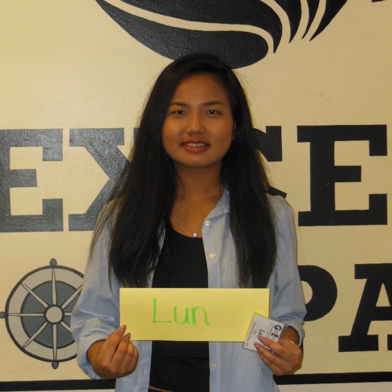 A person with long dark hair holds a yellow sign that says "Lun" while standing in front of a wall with black text and nautical symbols.