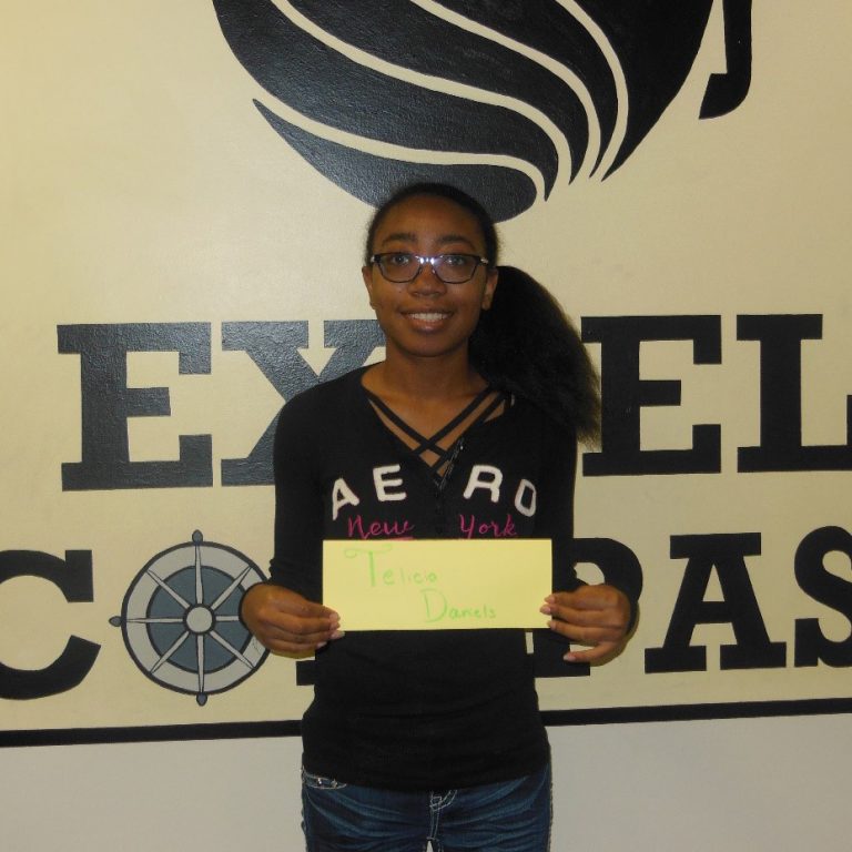A young person stands indoors holding a yellow sign with handwritten text in front of a wall with large letters and a compass symbol.