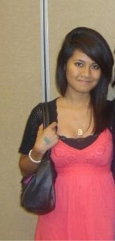 A woman with straight dark hair wearing a pink dress and black cardigan stands indoors, holding a black purse and smiling at the camera.