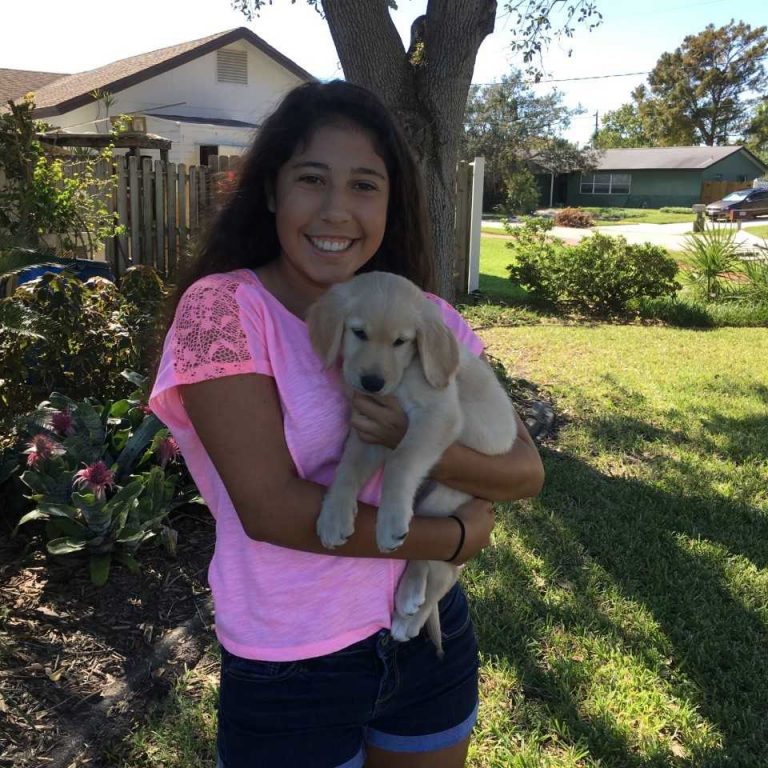 A young woman standing outside holds a light-colored puppy in her arms, smiling at the camera on a sunny day.