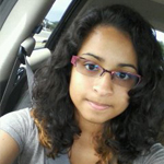 A young person with curly hair and glasses sits in a car seat, wearing a gray shirt and looking at the camera.