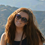 A woman with long brown hair wearing sunglasses and a black top stands outdoors with mountains in the background.