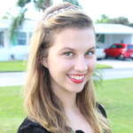 A young woman with long blonde hair and a braided headband smiles outdoors, standing in front of a green lawn, trees, and parked cars.