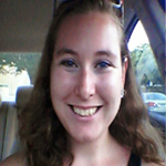 A person with long brown hair and sunglasses on their head, smiling while sitting in a car. Trees and parked cars are visible outside the window.