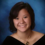 Young woman with short dark hair, wearing a black off-the-shoulder top and a necklace, poses smiling against a blue studio background.