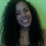 A woman with long curly hair is smiling at the camera in front of a light green background.