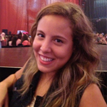 A woman with long wavy hair smiles at the camera while sitting in front of a stage at an indoor event.