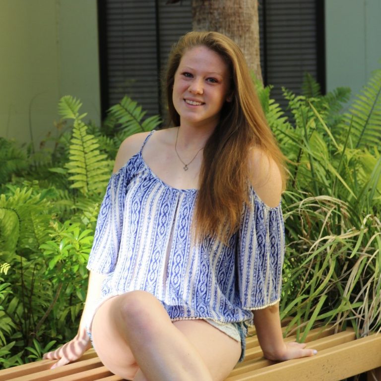 A young woman with long hair sits on a wooden bench outdoors, wearing a blue and white patterned top and shorts, surrounded by green plants.