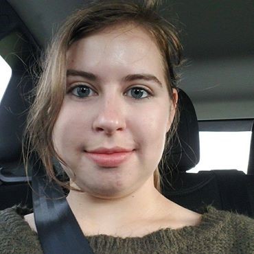 A young woman with light skin and brown hair sits in the back seat of a car, wearing a seatbelt and a green sweater, looking directly at the camera.