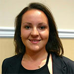A woman with shoulder-length dark hair is smiling at the camera. She is wearing a black blazer over a light top and standing in front of a beige wall with a white trim.