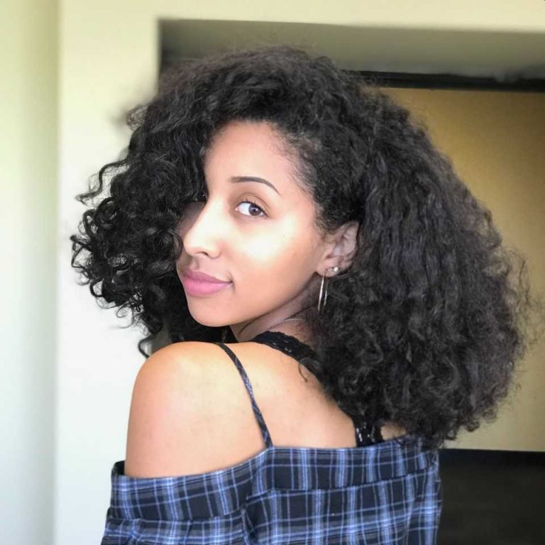 Woman with curly dark hair, wearing a blue plaid off-shoulder top and hoop earrings, looks over her shoulder in a well-lit indoor setting.