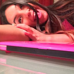 A woman lies on a table with her tongue out, playfully holding a strand of her hair, near a closed pink laptop.