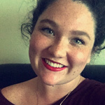 A person with curly dark hair, wearing red lipstick and a maroon top, smiling at the camera against a neutral background.