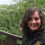 Woman with shoulder-length brown hair and a black jacket sitting outdoors on a wooden deck, with green trees in the background.