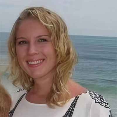 A young woman with blonde wavy hair smiles at the camera, standing in front of a calm ocean under a cloudy sky.