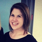 A woman with shoulder-length brown hair smiles at the camera while standing indoors, wearing a black top.