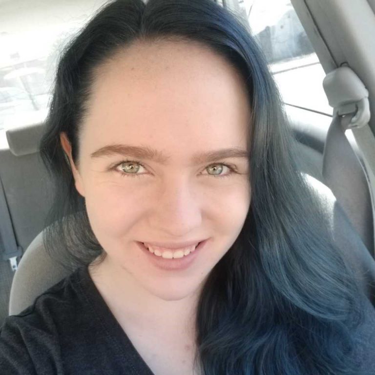 A person with long dark hair and light eyes smiles at the camera while sitting in a car.